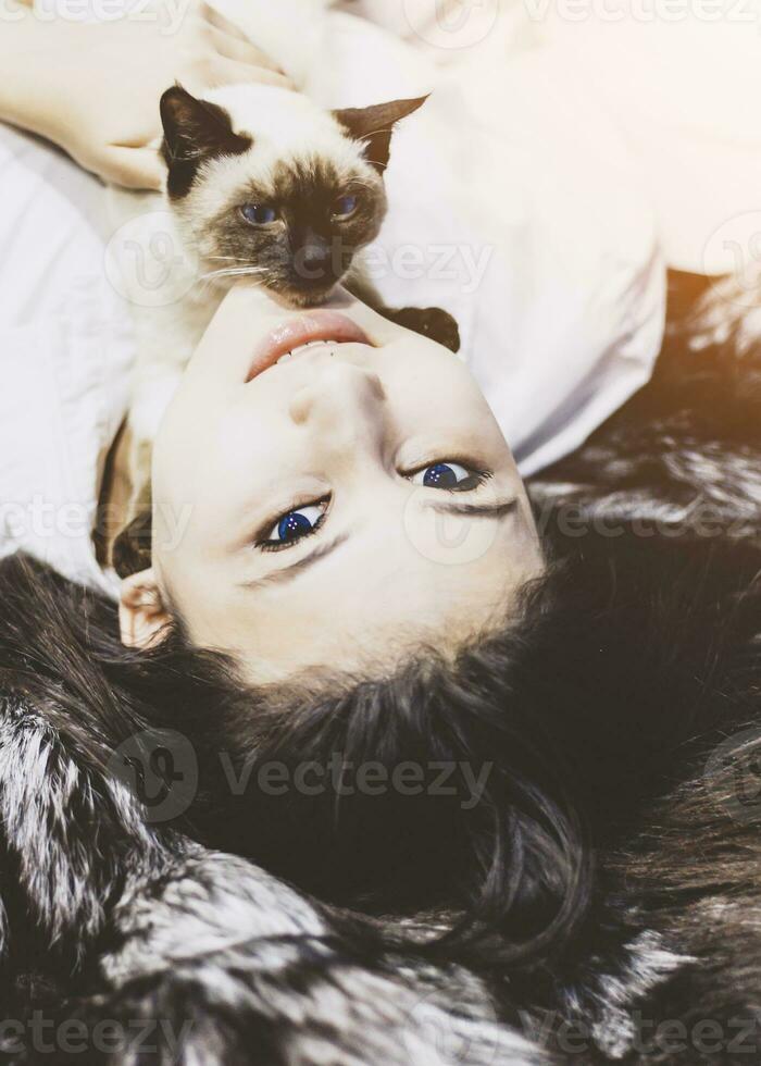 Young middle-eastern woman lying on bed and having fun with her Siamese cat. Pet loving concept. View from above to cute girl playing with cute kitten photo