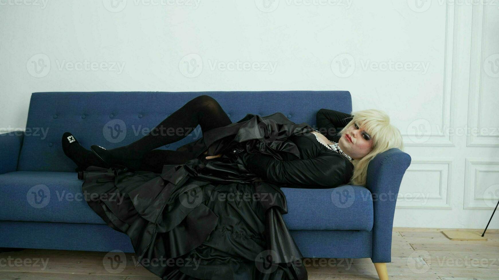 A drag queen in a black dress lies on a sofa. A drag queen in white wig and black evening dress. photo