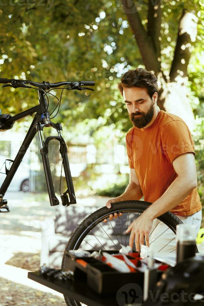 In home yard, young energetic man is using expert tools to replace damaged bike wheel. Servicing on disassembled bicycle outside for pleasure cycling is a committed, healthy caucasian male cyclist. photo
