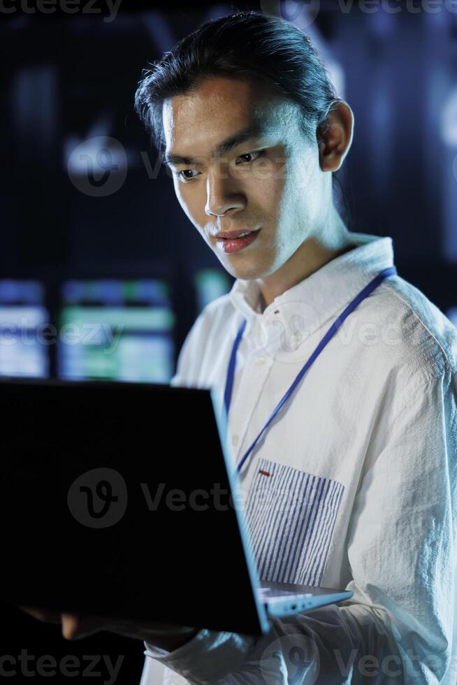Dara center knowledgeable professional running code and programming on laptop, troubleshooting and upgrading high tech facility designed to accommodate rows of fully operational server electronics photo