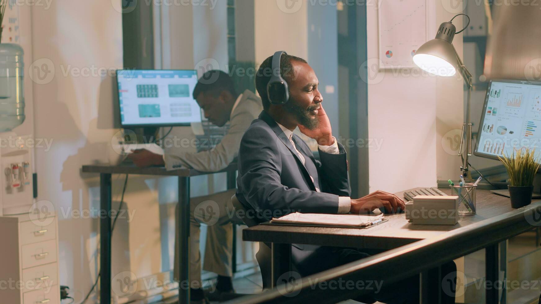 africano americano trabajador disfrutando música a tomar su mente lejos desde aburrido rutina proyecto Tareas durante Turno nocturno. empresario en oficina con auriculares en comparando márketing gráficos y estadísticas foto