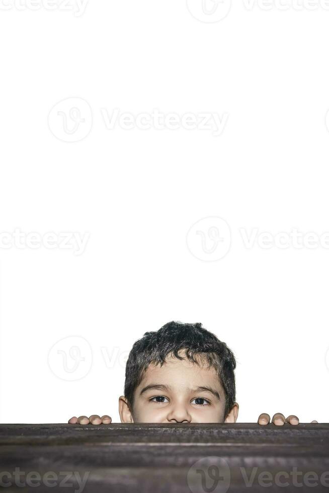A kid peeking out from under the table. Cute young mixed race 4-5 years old boy looking over wooden desk on white background with copy space photo