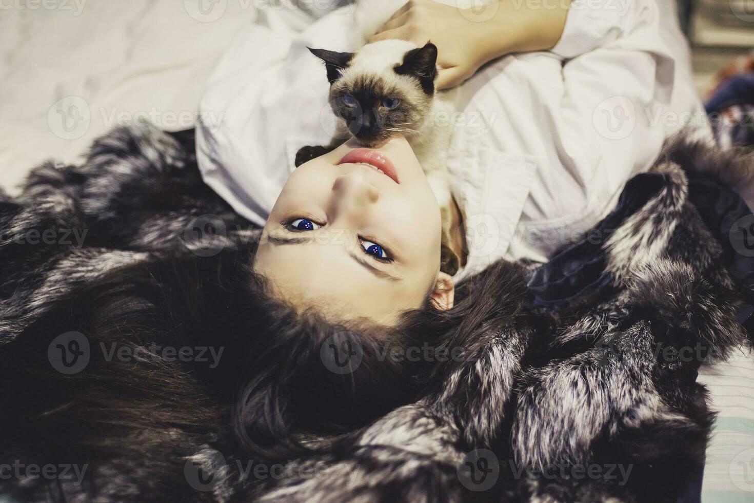 Young middle-eastern woman lying on bed and having fun with her Siamese cat. Pet loving concept. View from above to cute girl playing with cute kitten photo