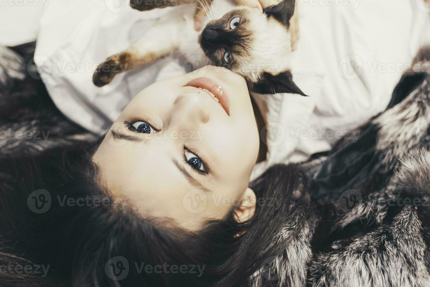 Young middle-eastern woman lying on bed and having fun with her Siamese cat. Pet loving concept. View from above to cute girl playing with cute kitten photo