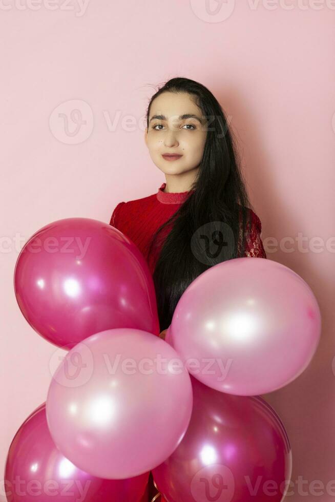 Young pretty woman in red evening dress holding festive air balloons. Portrait of happy 20s middle-eastern female celebrating valentines day, birthday, party photo