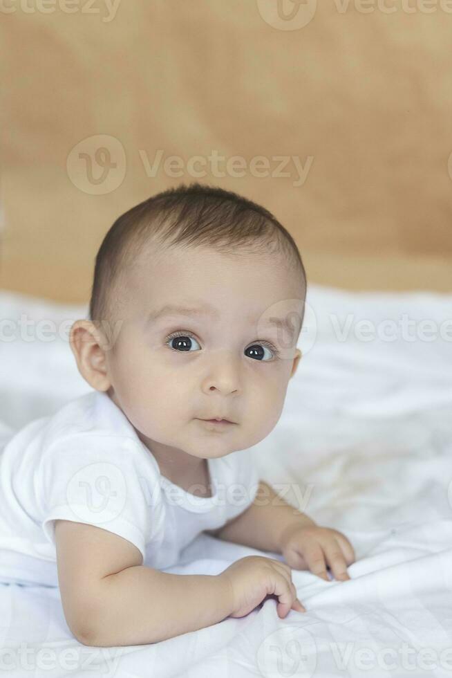 6-8-month-old baby boy lying playfully in bed. Charming 6-7 month little baby in white bodysuit. Baby boy in white bedding. Copy space photo