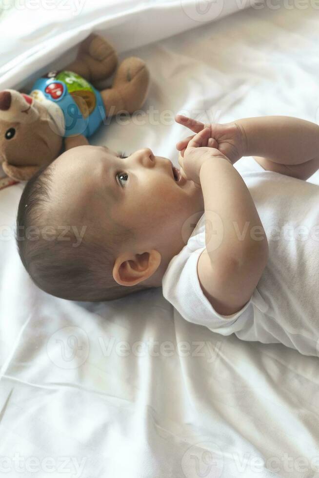6-8 meses bebé chico acostado en broma en cama. encantador 6-7 mes pequeño bebé en blanco mono bebé chico en blanco lecho. Copiar espacio foto