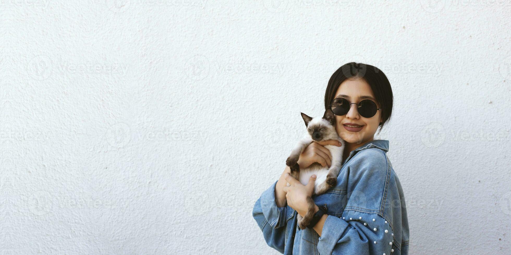 joven atractivo mujer abrazando coño gato en manos. linda y atractivo niña en de moda Gafas de sol posando con su siamés gato foto