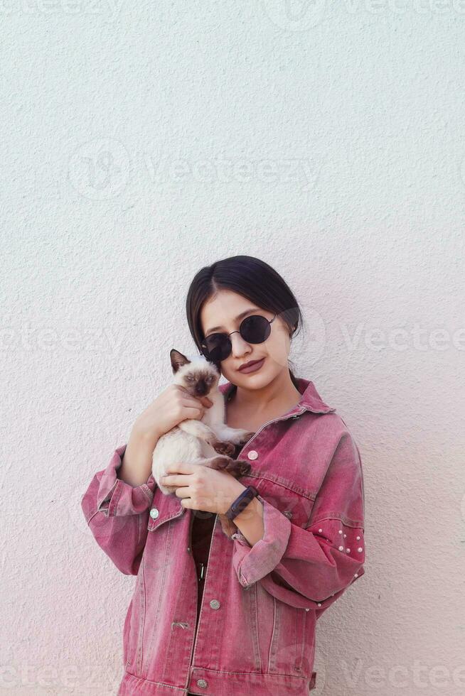 joven atractivo mujer abrazando coño gato en manos. linda y atractivo niña en de moda Gafas de sol posando con su siamés gato foto