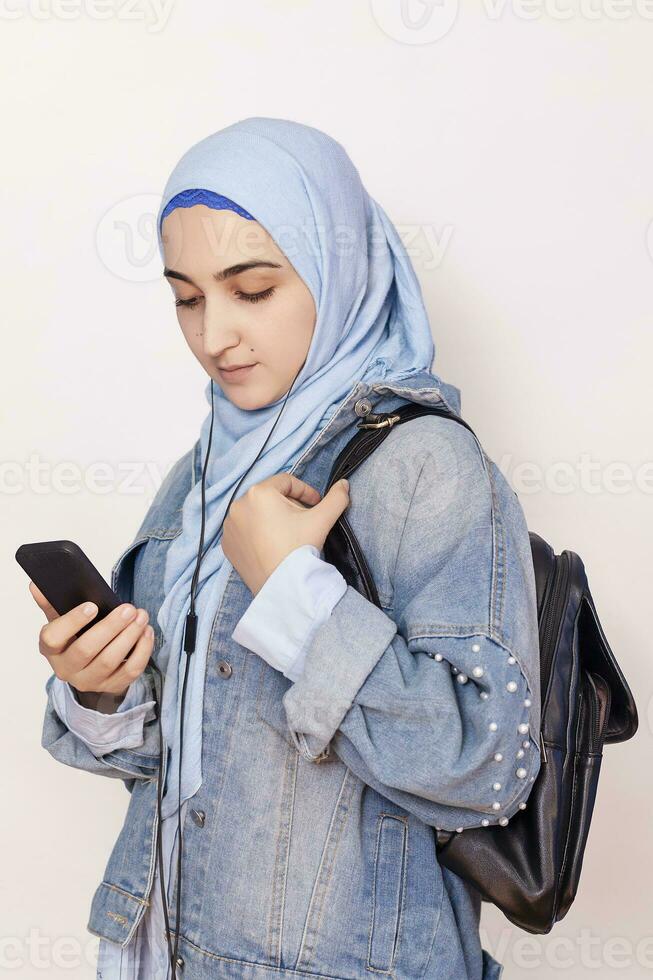 moderno musulmán estudiante niña escucha a música, podcasts, y audiolibros en el teléfono inteligente contento musulmán mujer en hijab escuchando canciones con el auricular foto