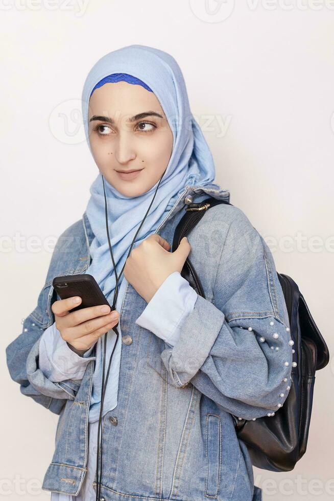 moderno musulmán estudiante niña escucha a música, podcasts, y audiolibros en el teléfono inteligente contento musulmán mujer en hijab escuchando canciones con el auricular foto