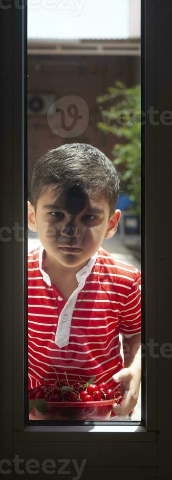 Little boy standing outside with a bowl cherries and looking through the door. Childhood, summertime concept photo