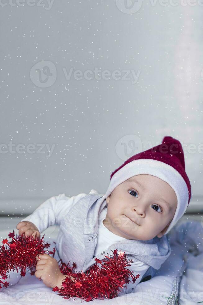 Little funny Santa boy playing with christmas or new year holiday festive garland. 1 year old baby boy in Santa Claus cap. photo