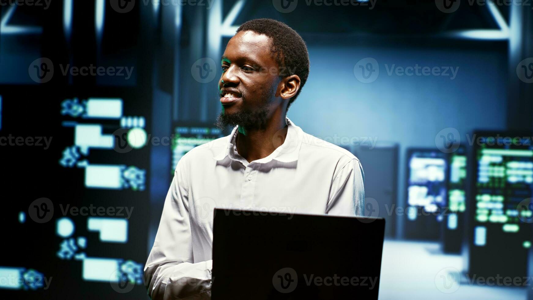 Happy african american employee using laptop to review energy consumption across server cabinets components. Worker making sure data center electronics temperature sensors are operating perfectly photo