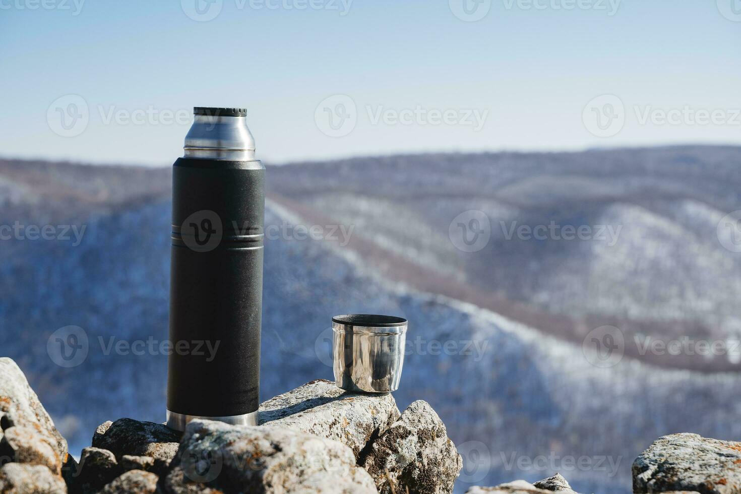 A cup of hot coffee against the sky. Thermos for tea, Drink in a black bottle stands on the stones. The concept of dishes for tourism, the equipment of the traveler. photo