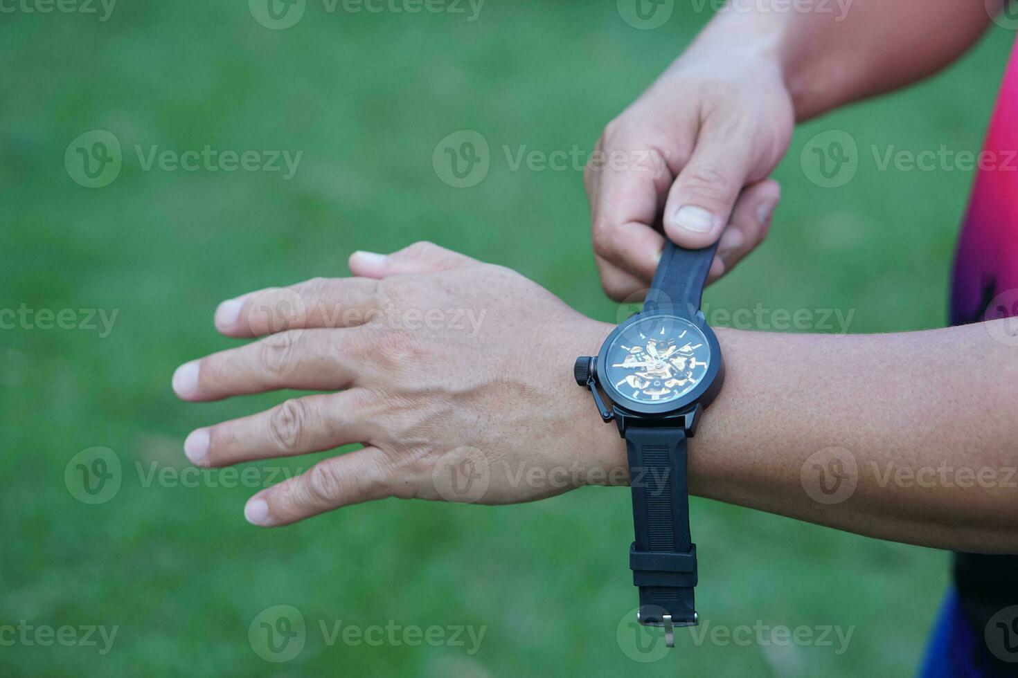 Close up man hand is wearing black watch on his wrist, green outdoor background. Concept, fashionable accessory for time telling, make you look stylish, punctual and smart. Analog wrist watch. photo