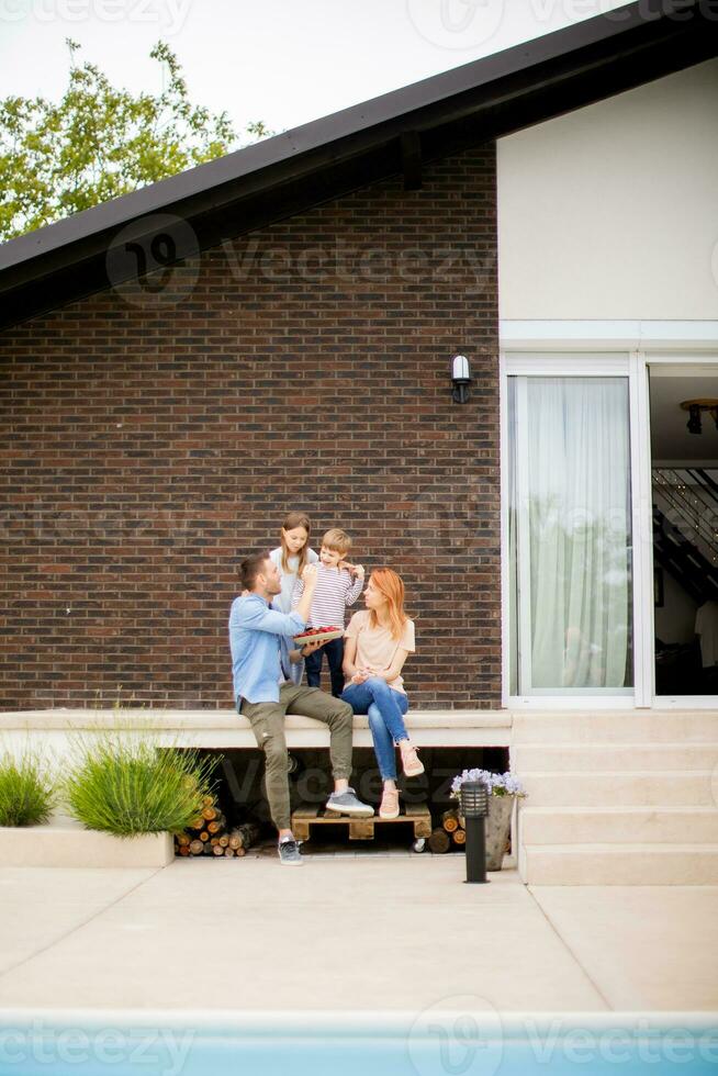 Family with a mother, father, son and daughter sitting outside on steps of a front porch of a brick house and eating strawberries photo