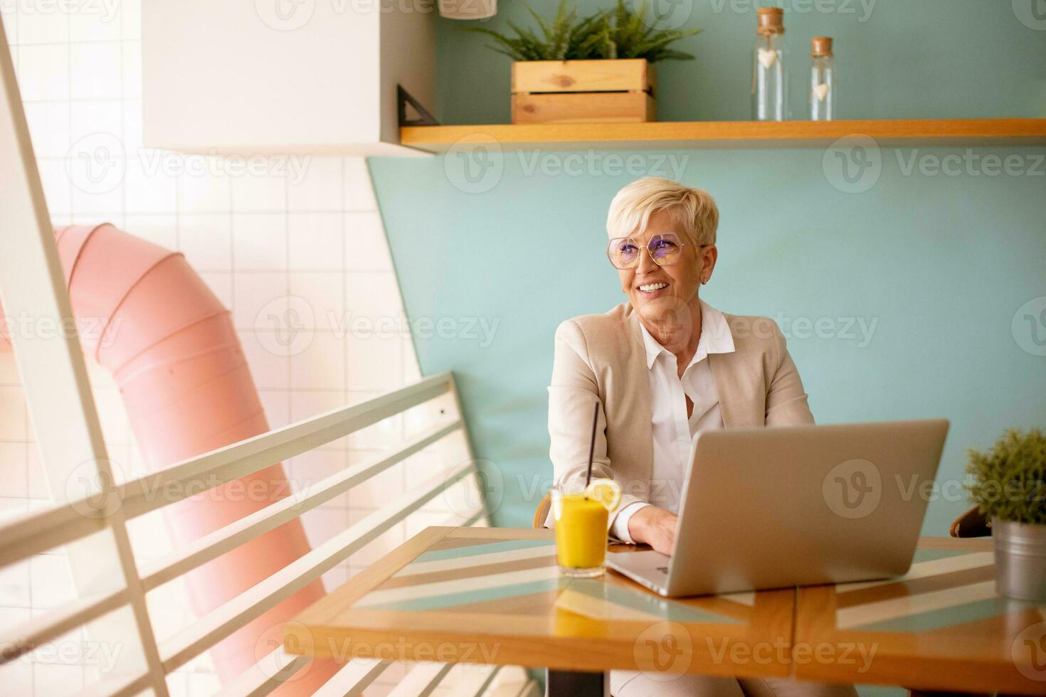 Senior woman using mobile phone while working on laptop and drinking fresh orange juice in the cafe photo
