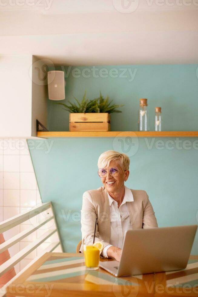 Senior woman using mobile phone while working on laptop and drinking fresh orange juice in the cafe photo