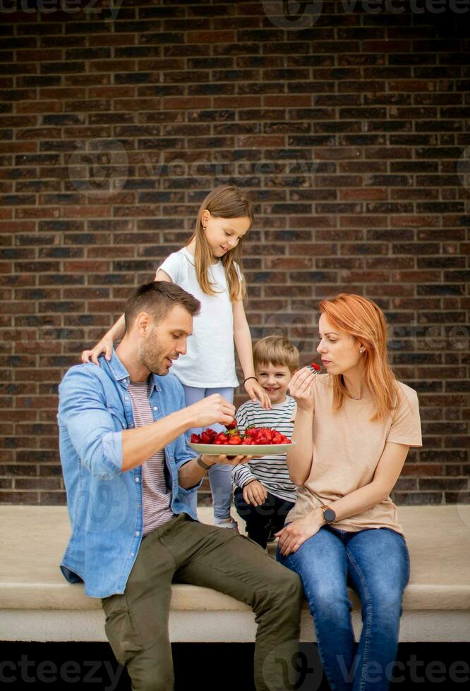 Family with a mother, father, son and daughter sitting outside on steps of a front porch of a brick house and eating strawberries photo