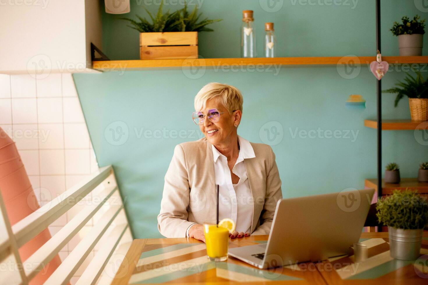 Senior woman using mobile phone while working on laptop and drinking fresh orange juice in the cafe photo
