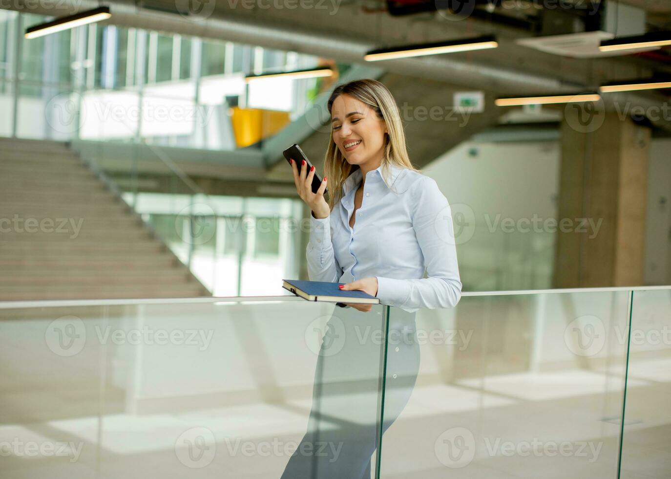 Young woman with notebook using mobile phone and standing in the modern office hallway photo