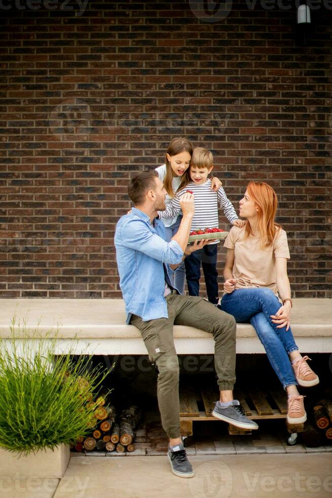 Family with a mother, father, son and daughter sitting outside on steps of a front porch of a brick house and eating strawberries photo