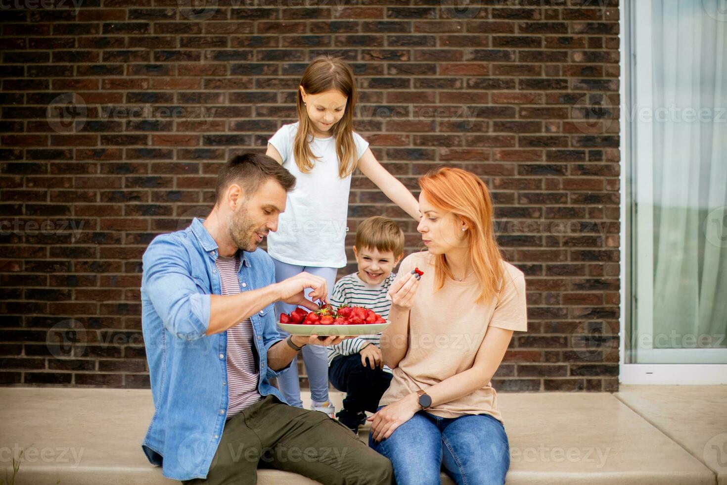 Family with a mother, father, son and daughter sitting outside on steps of a front porch of a brick house and eating strawberries photo