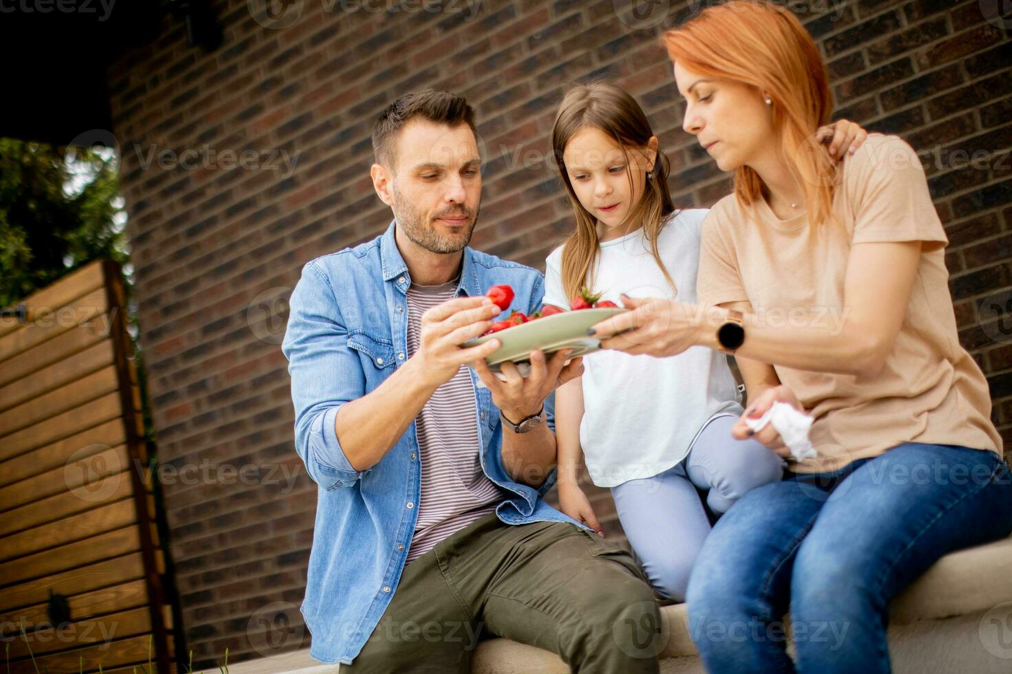 Family with a mother, father and daughter sitting outside on steps of a front porch of a brick house and eating strawberries photo