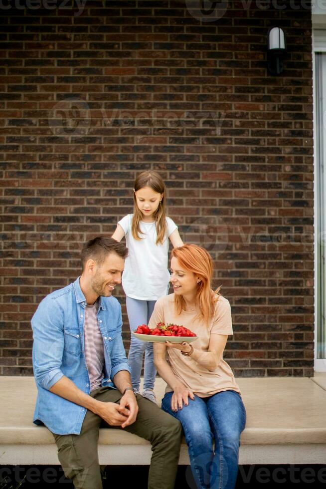 Family with a mother, father and daughter sitting outside on steps of a front porch of a brick house and eating strawberries photo