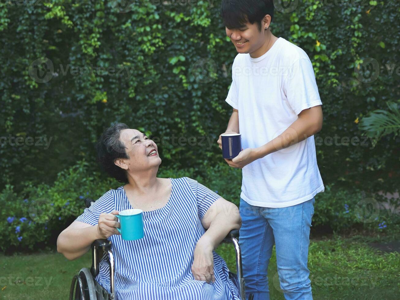 asiático mayor mujer sentado en silla de ruedas, Bebiendo café o té con su hijo en el jardín. sonriente felizmente. foto