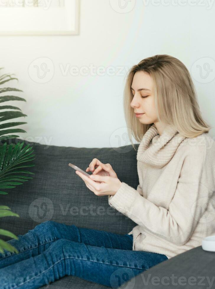 Happy young woman relaxing on comfortable couch, holding smartphone in hands. photo