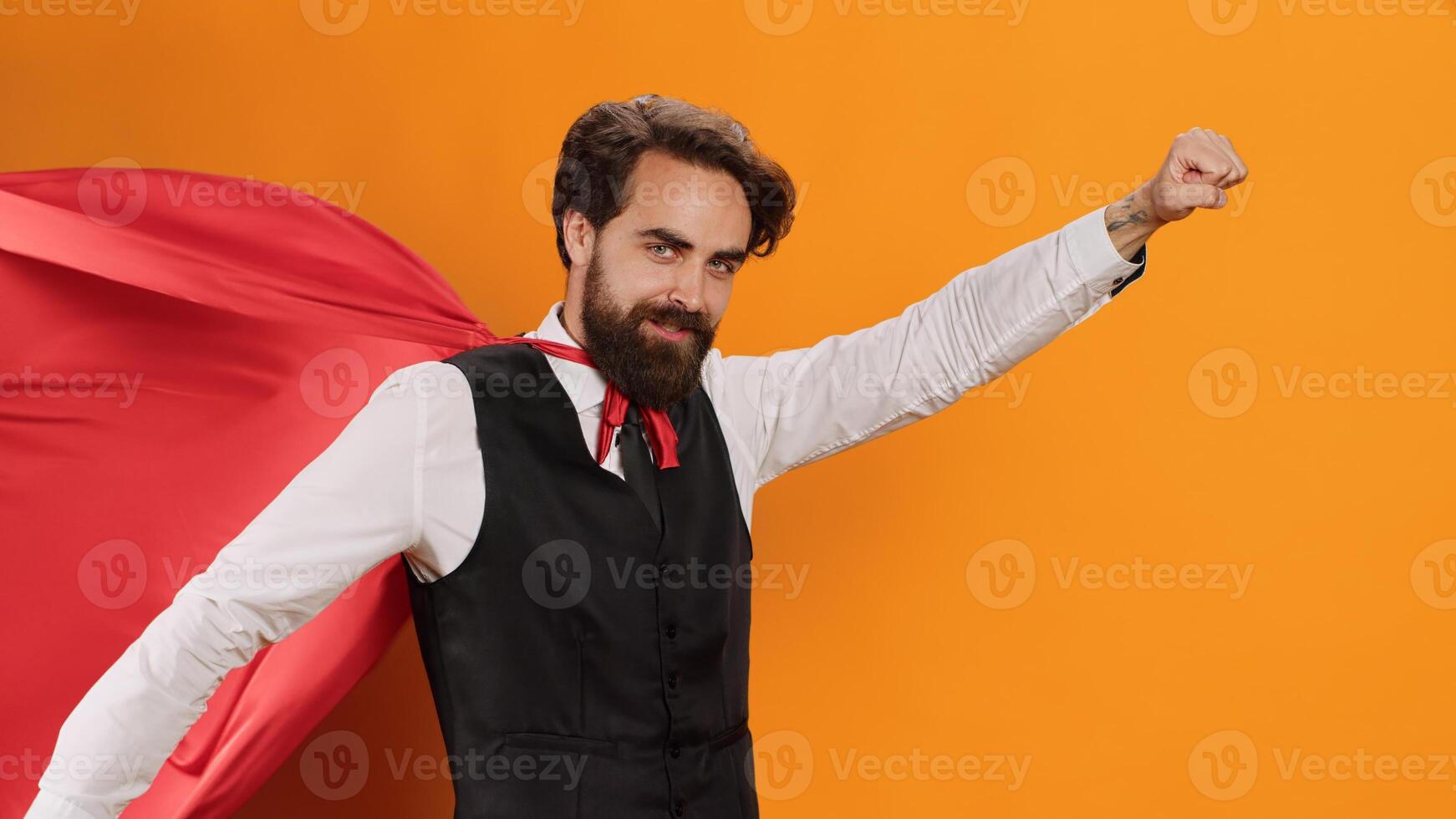 Elegant butler with red cape posing on camera while he acts like superhuman at luxury restaurant, yellow studio. Young man catering worker serving food and drinks, wearing hero mantle. photo
