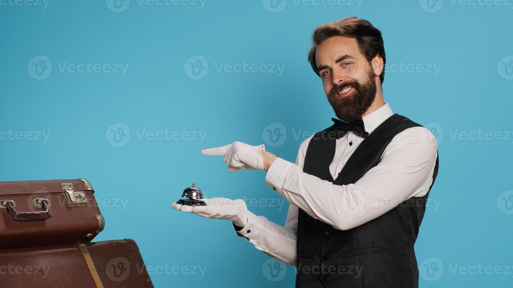 Hotel porter points to service bell, advertising concierge assistance and ringing to provide luxury service with baggage. Young professional doorman helping with trolley bags, hospitality industry. photo