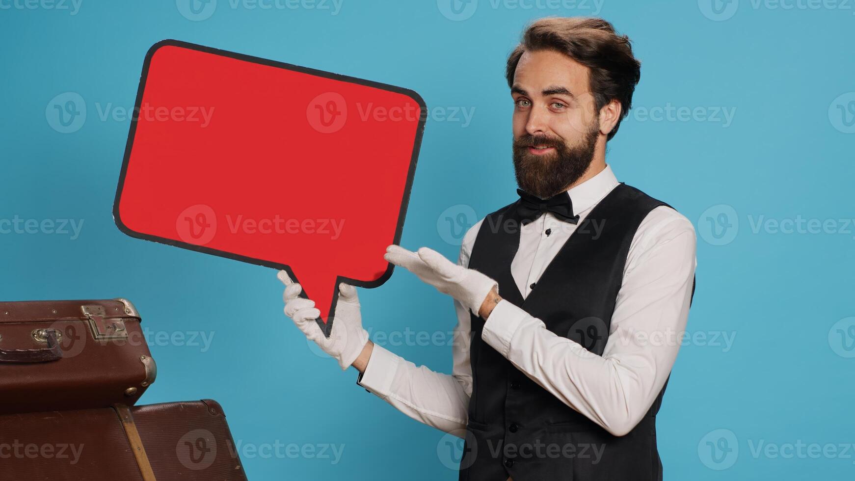 Hotel concierge shows ad with speech bubble, indicating empty isolated red cardboard icon. Skilled bellboy presents blank copyspace billboard sign in studio against blue background. photo