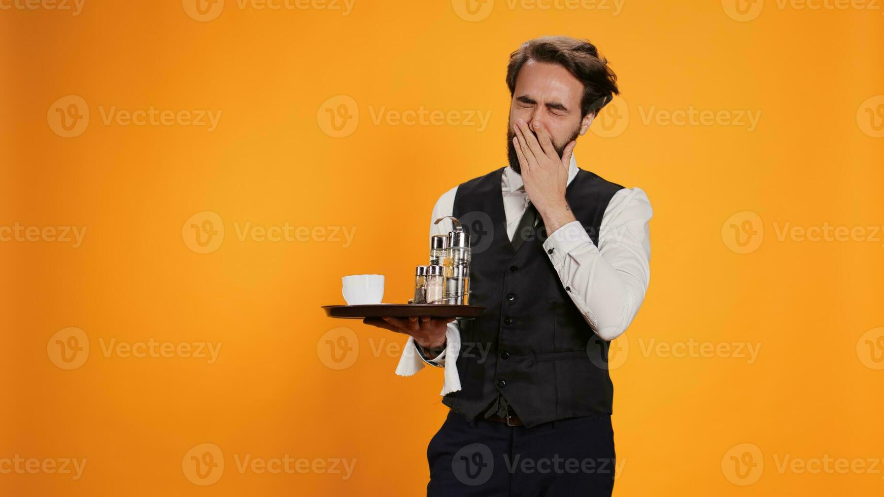 Exhausted butler feeling sleepy on camera, yawning and falling asleep on duty at restaurant. Waiter in formal attire feeling tired and stretching in studio, standing against yellow background. photo