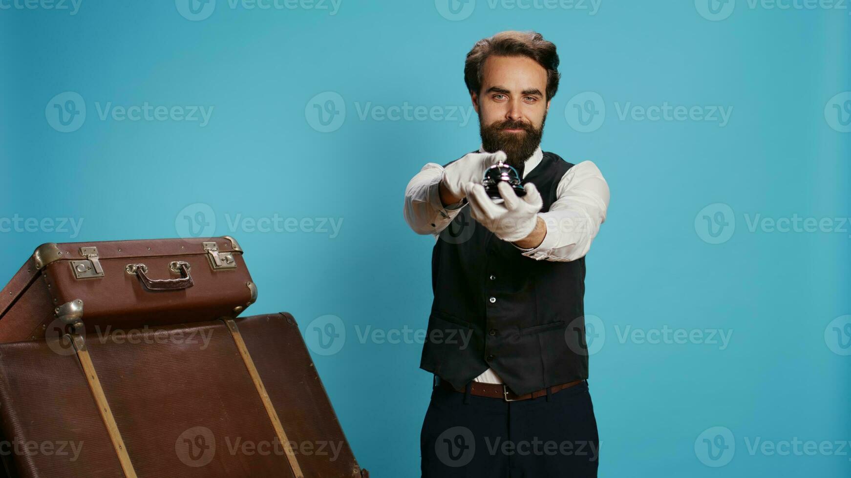 Stylish bellboy with luggage and bell, using to ring for concierge service at hotel in tourism sector. Doorkeeper porter handling trolley bags to help guests, classy elegant occupation. photo