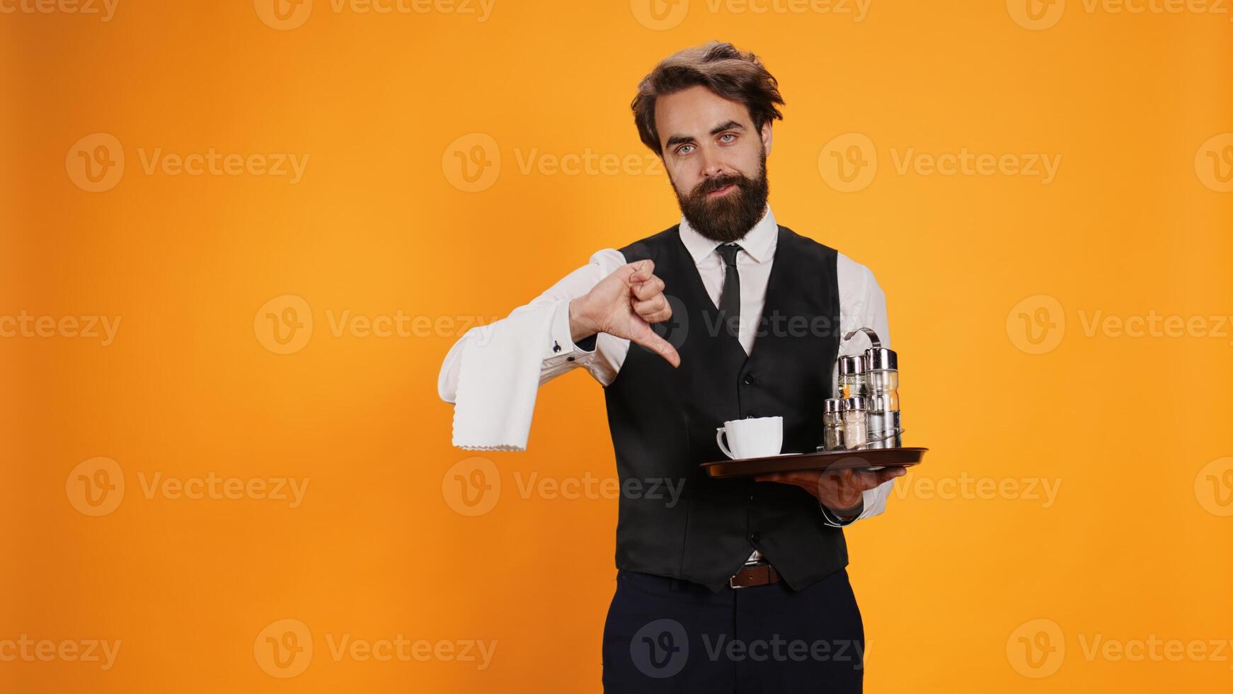Staff member gives thumbs down sign on camera, showcasing dislike and disagreement sign against yellow background. Skilled waiter presents disapproval symbol, carrying restaurant food platter. photo