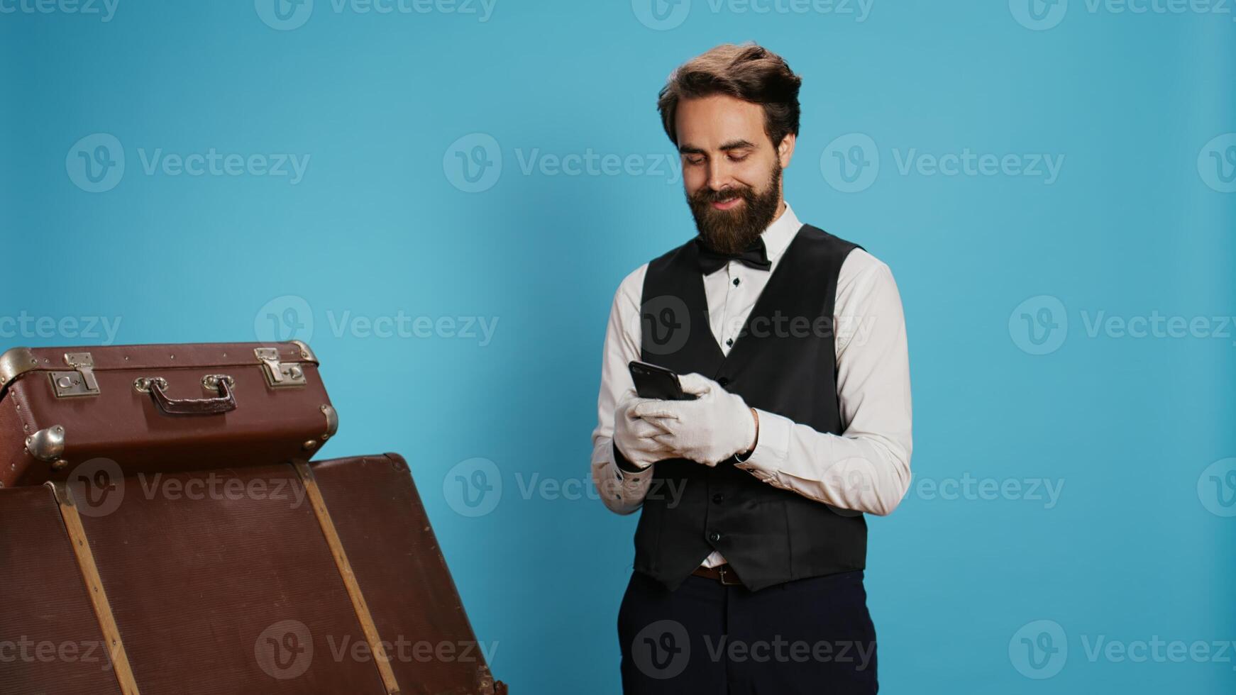 Doorkeeper browses social media app on camera, using smartphone to text messages or review list of online reservations. Professional hotel porter texting on mobile phone, studio shot. photo