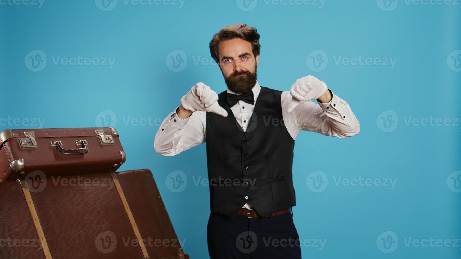 Staff member being dissatisfied giving thumbs down on camera, standing next to trolley bags. Experienced bellboy showcases negative feelings of rejection, expressing disappointment. photo