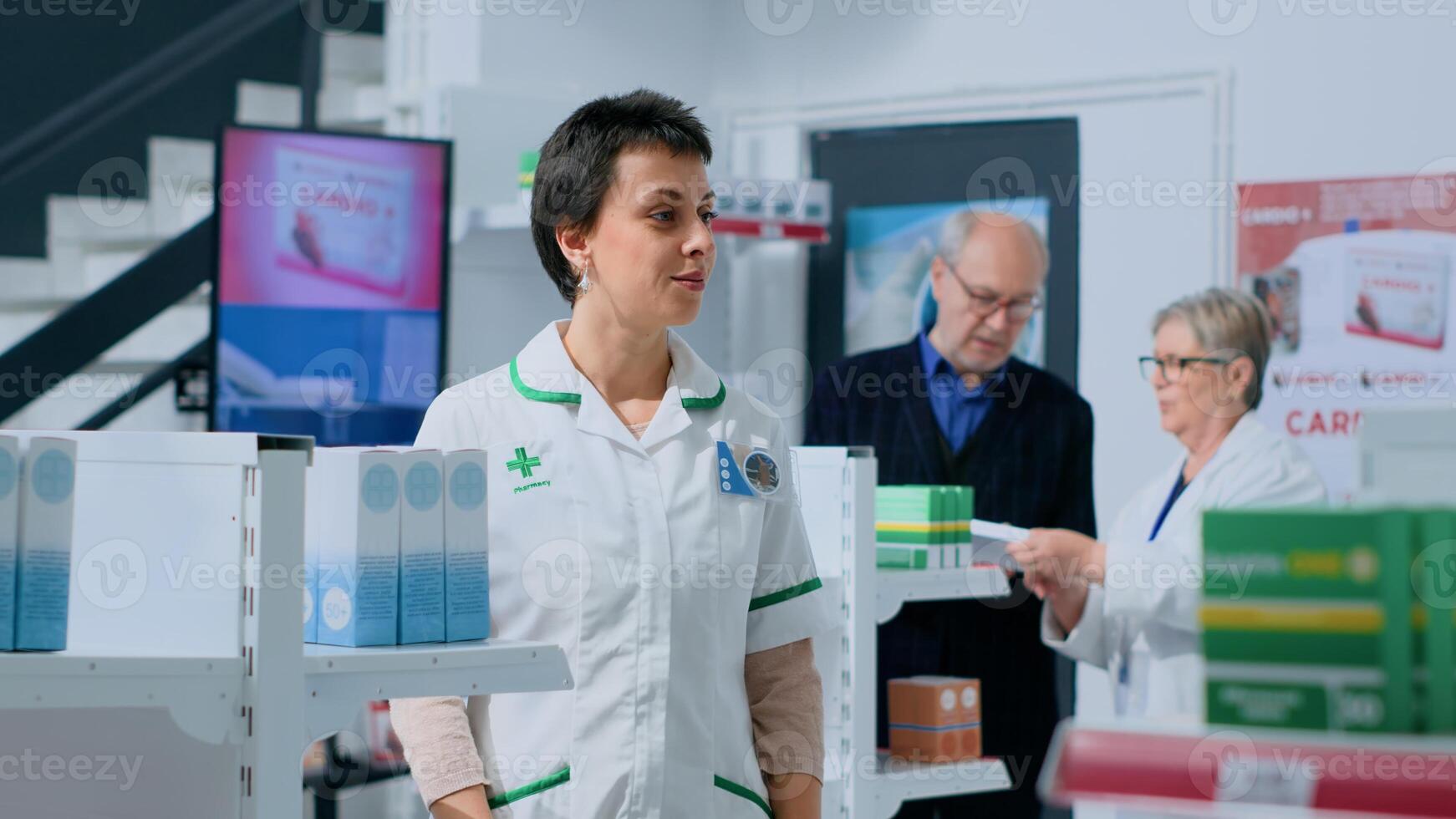 Friendly pharmacist in apothecary placing pills box on shelves while elderly coworker helps customer with informations. Smiling professional in drugstore arranging medicinal products photo