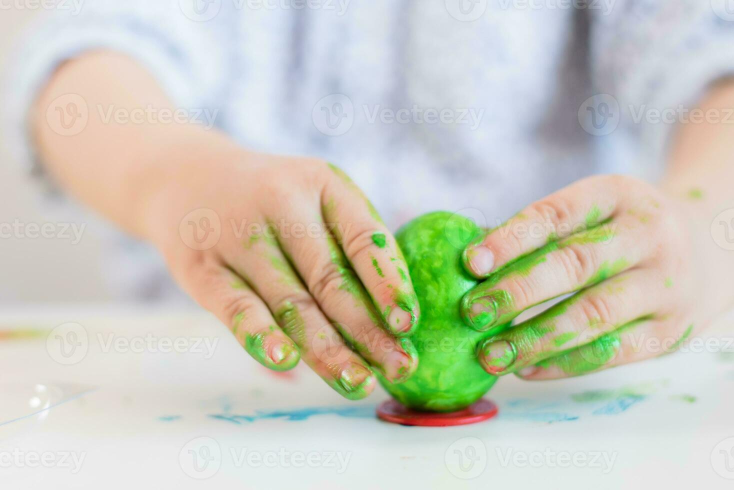 un niño pone un verde Pascua de Resurrección huevo en un estar con su manos manchado con pintar en un blanco mesa. foto