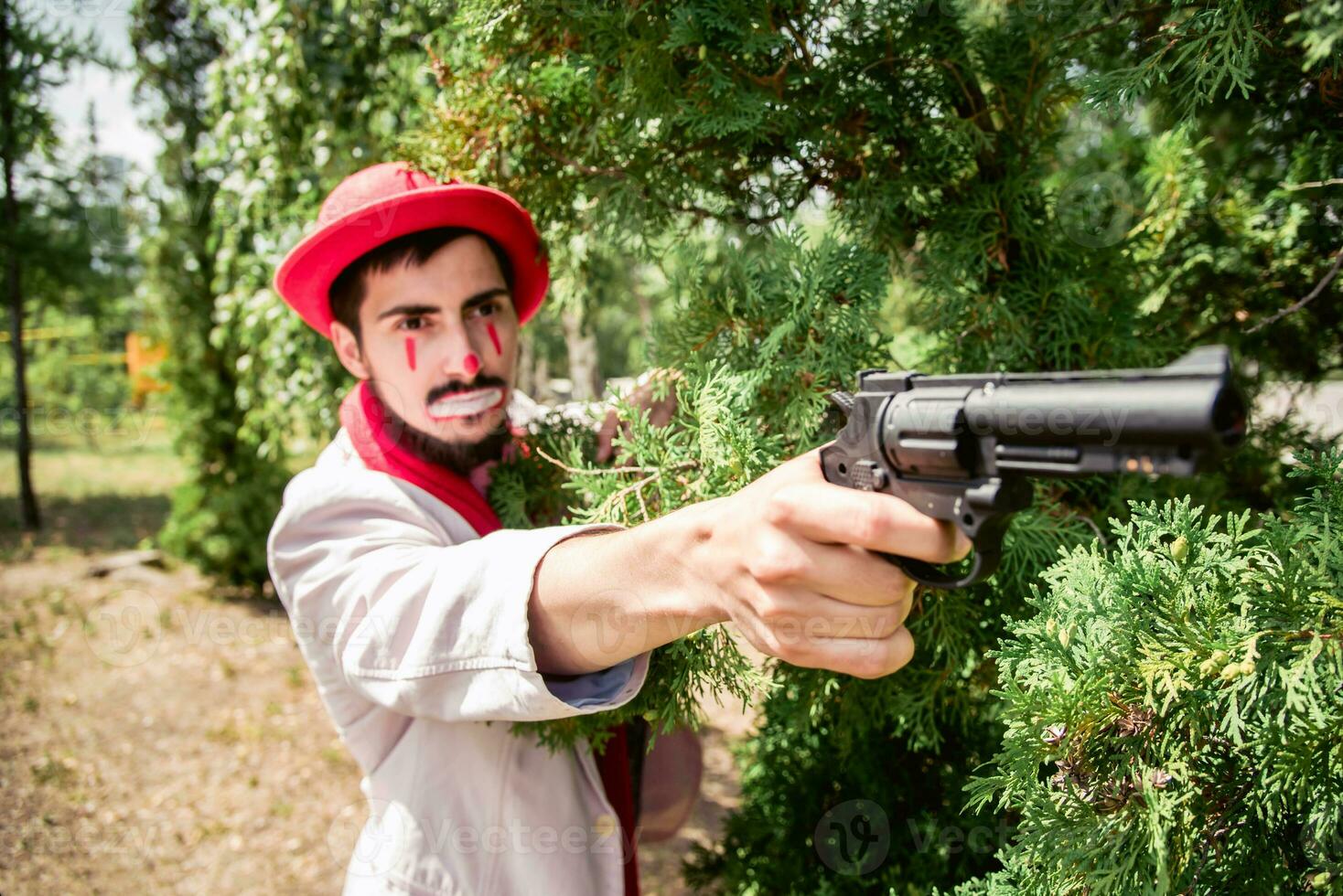 Mime in the guise of a villain peeks out from behind a tree and threatens with a pistol. Evil clown with a gun in the park. photo