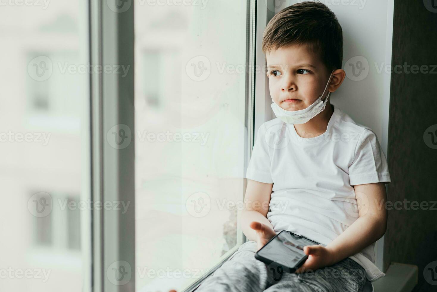 A small child in a medical mask sits quarantined at home on a window with a phone in his hands.Prevention of coronavirus and Covid - 19 photo