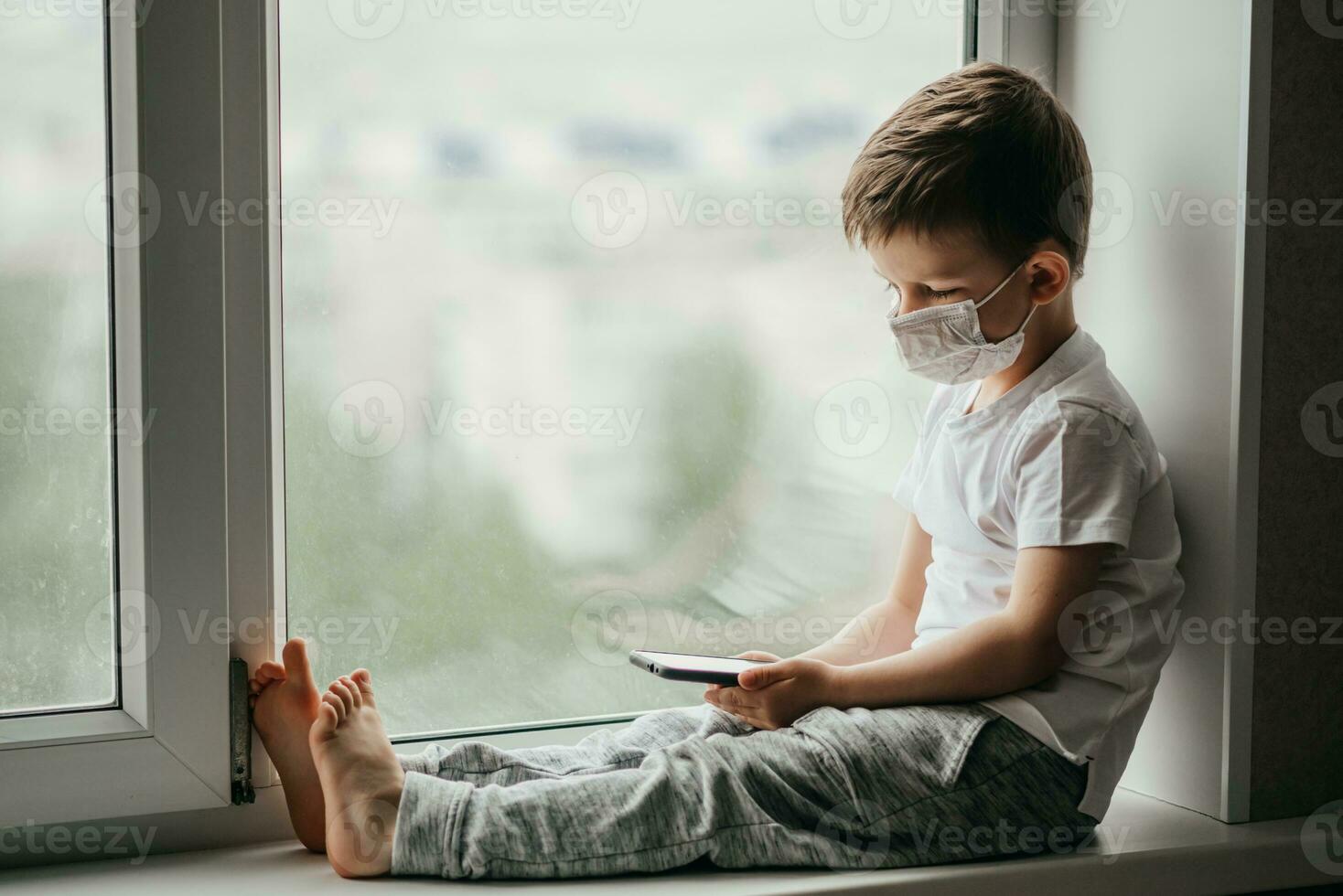 A small child in a medical mask sits quarantined at home on a window with a phone in his hands.Prevention of coronavirus and Covid - 19 photo