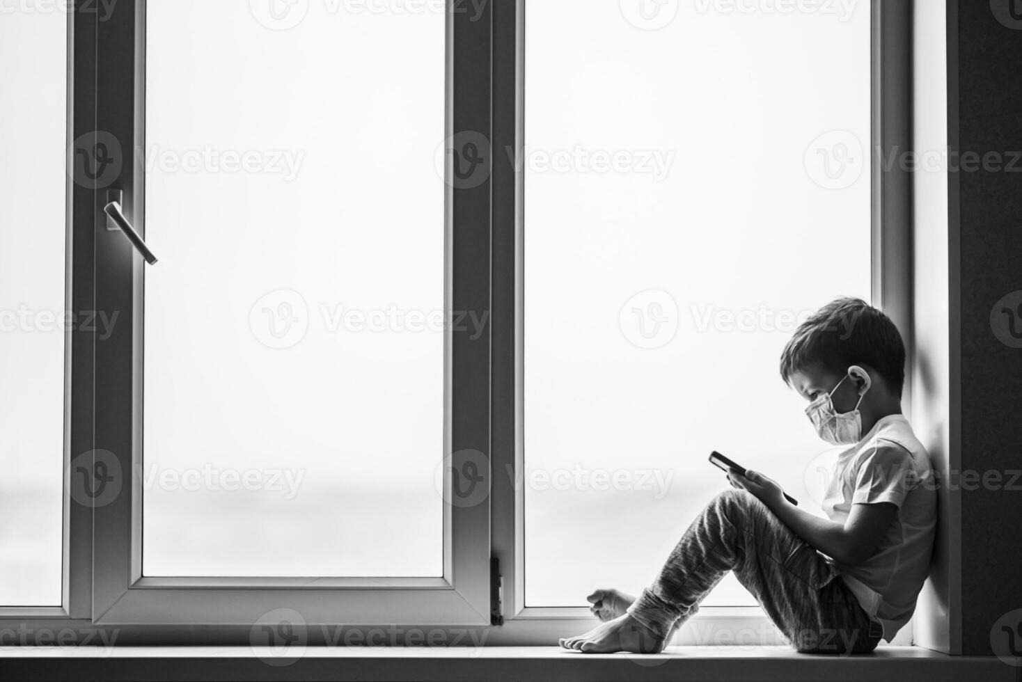 A small child in a medical mask sits quarantined at home on a window with a phone in his hands.Prevention of coronavirus and Covid - 19 photo