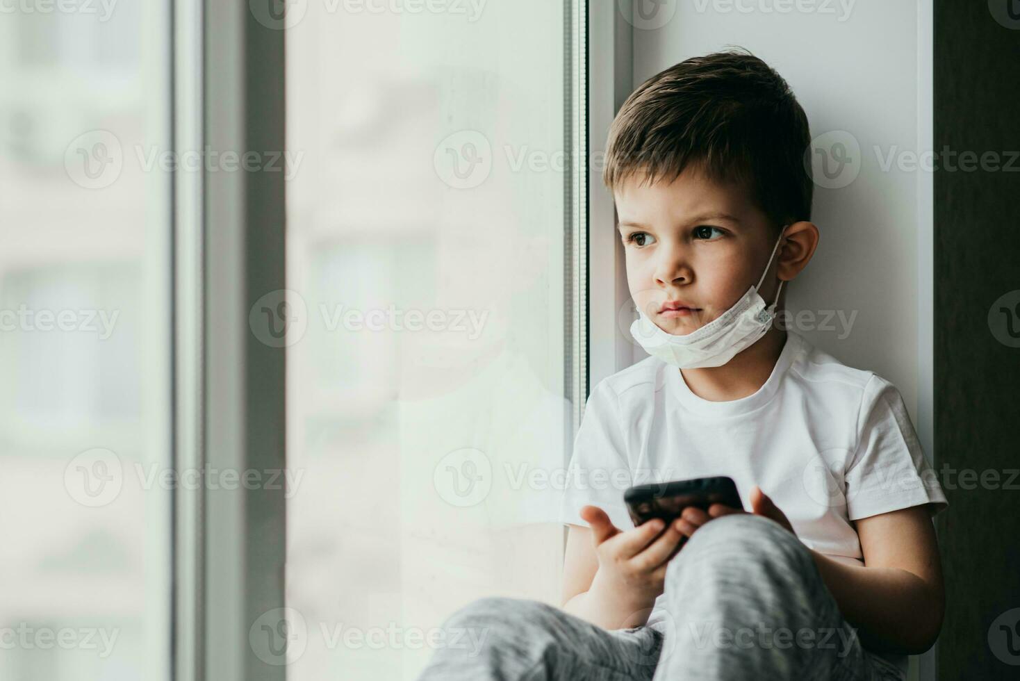 A little boy in a medical mask is sitting on the window of the house in quarantine with a phone in his hands.Prevention of coronavirus and Covid - 19 photo