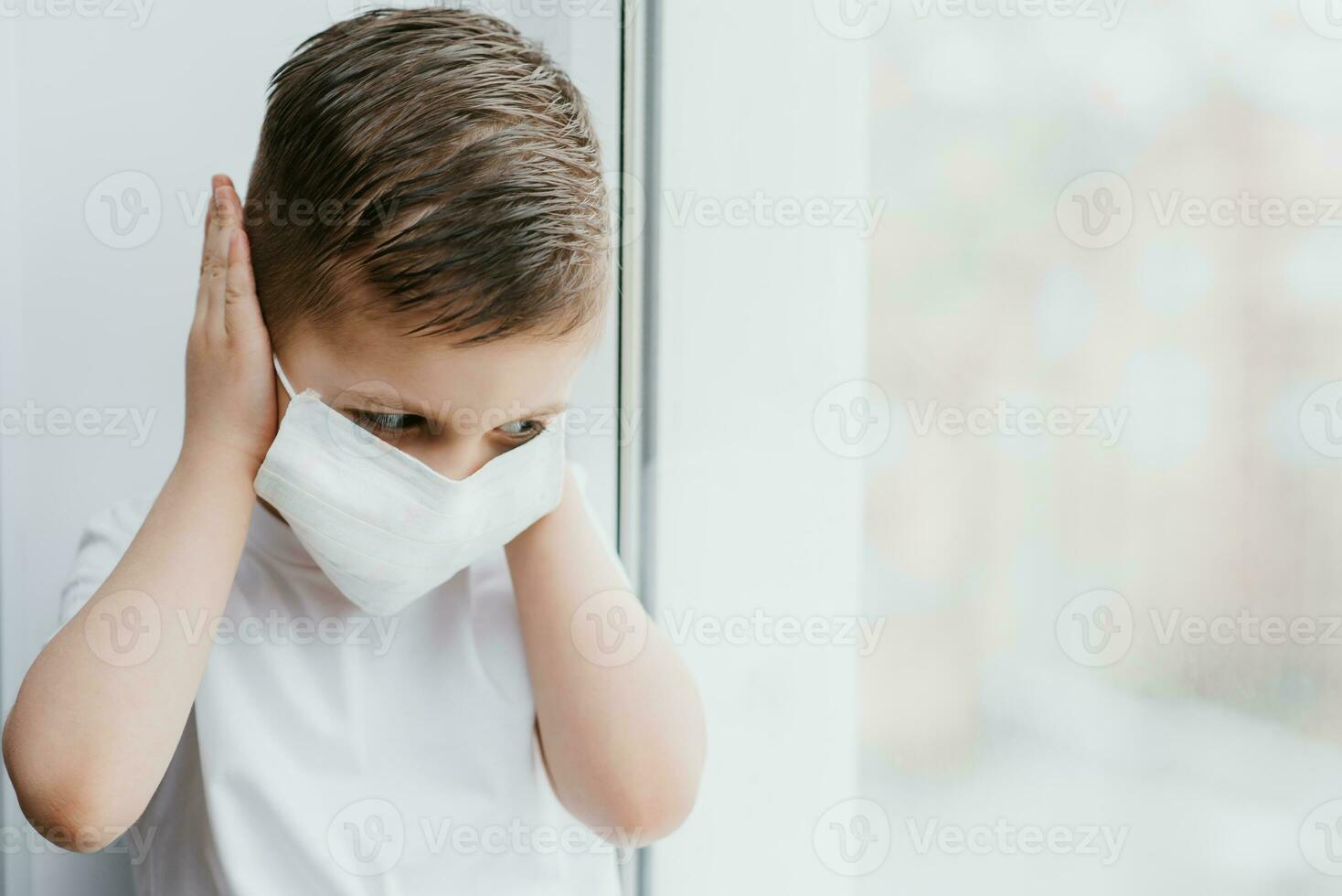 a child in a medical mask is sitting at home in quarantine because of coronavirus and covid -19 and looks out the window. photo