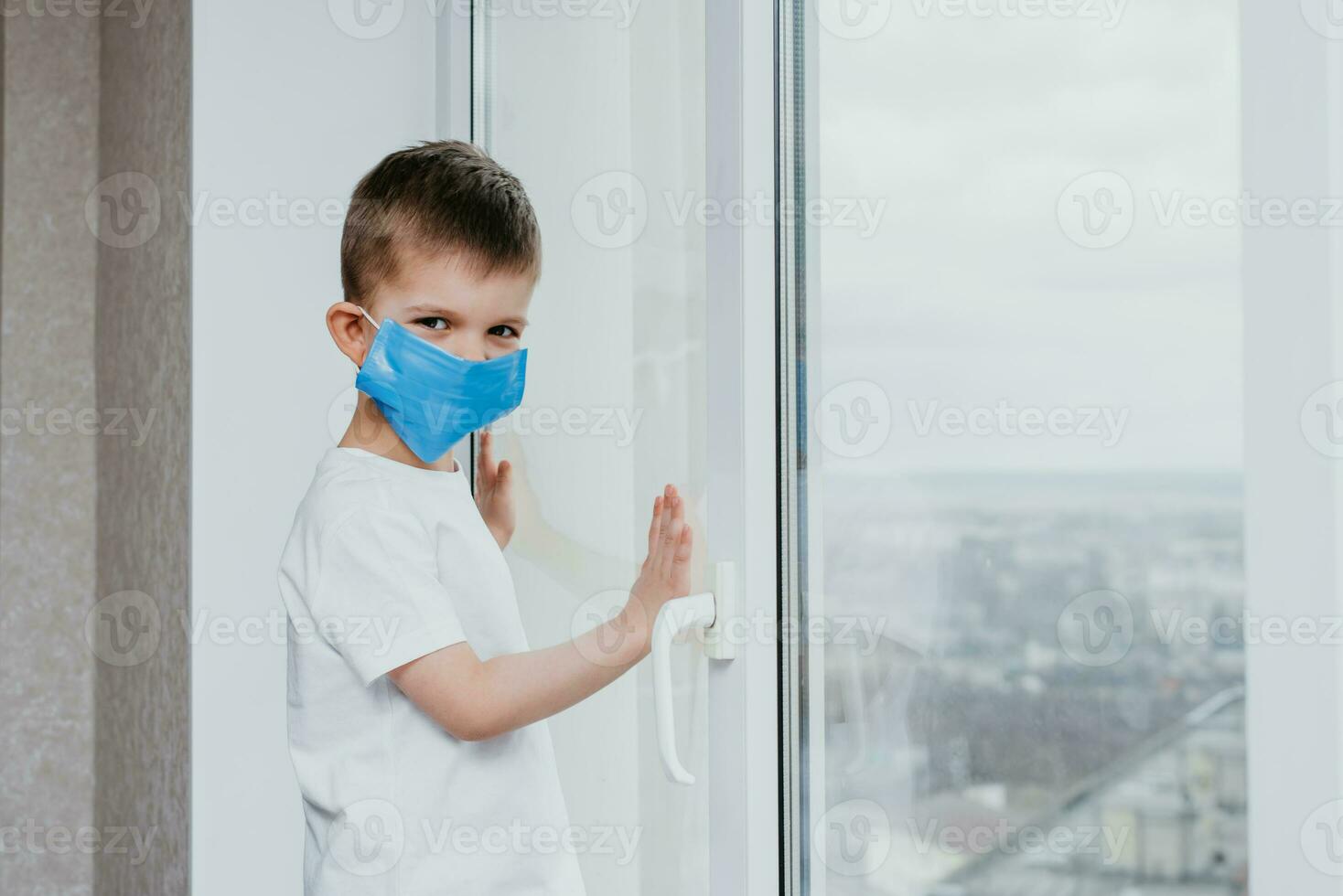 a child in a medical mask is sitting at home in quarantine because of coronavirus and covid -19 and looks out the window. photo