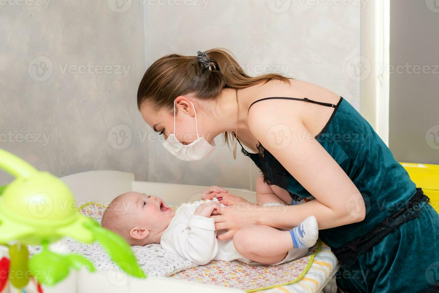 A mother in a medical protective mask takes care of her baby at home in a corontine. Prophylactic protection of coronavirus and covid-19 during a pandemic. photo